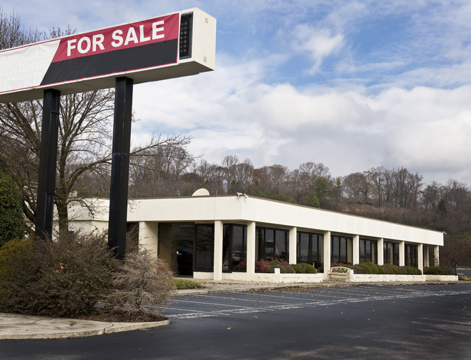 Vacant commercial building with 'For Sale' sign above, clear sky background.