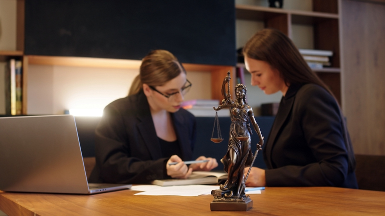 Two women in a law office discussing documents with Lady Justice statue.
