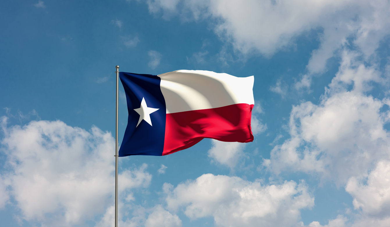 Texas state flag waving against a clear blue sky with clouds.