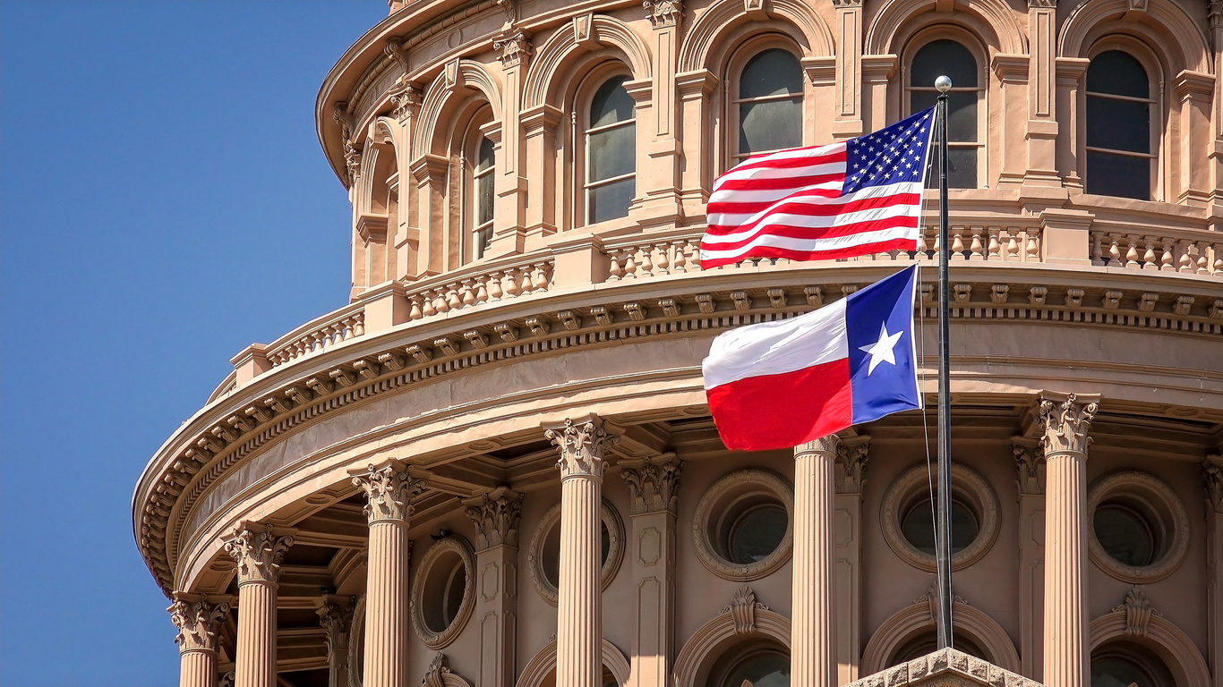 Texas and American flags flying in front of the Texas Capitol building dome.