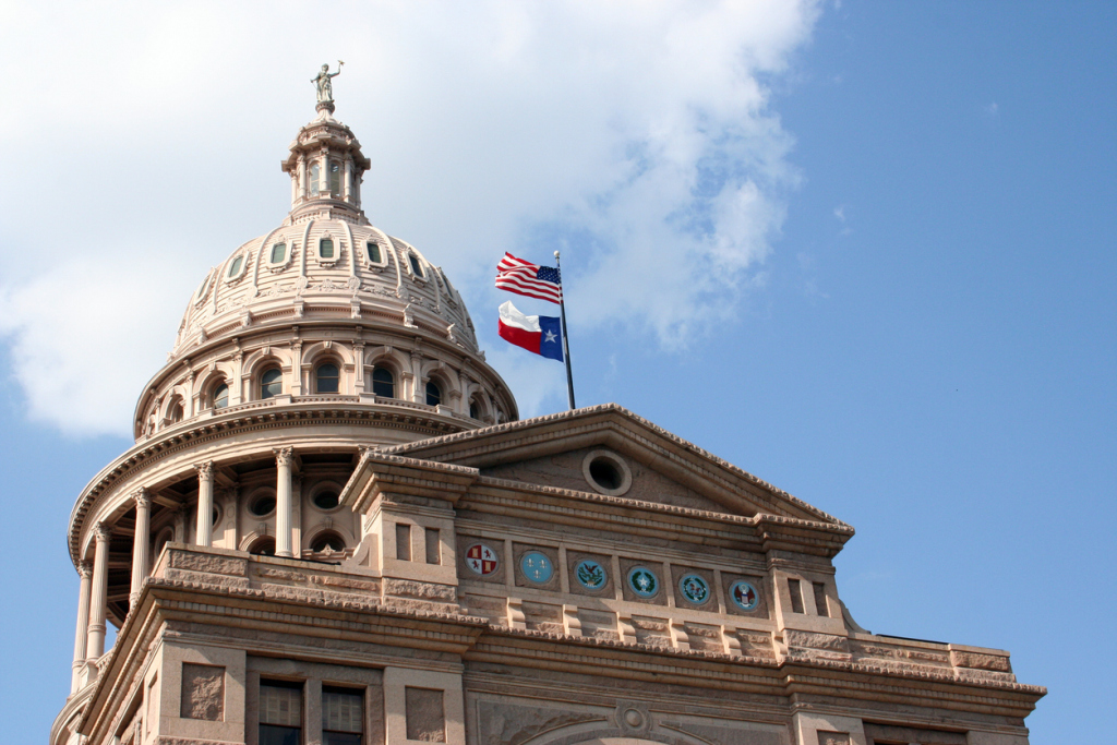 Texas State Capitol dome with American and Texas flags against blue sky.